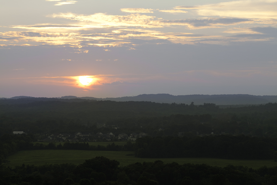 This Brian Charles Steel photo depicts a landscape at sunset in Cleveland, Tennessee.  The sun is towards the middle portion of the frame on the right side of the image.  Below the sun are green mountains covered with trees, and a grass plain. The plain is towards the bottom of the image, and below it in the foreground are more trees.  Just above the plain on the right side is a large cluster of houses on the side of a mountain.  The sky occupies the top half of the frame the bottom portion of the clouds are bluish mixed with streaks of orange from the sun.  The clouds above that portion of the sky are orange and yellow with a few blue streaks.  